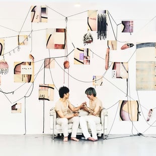 ソファに座る男性二人と白い壁に飾られた多数のアート作品