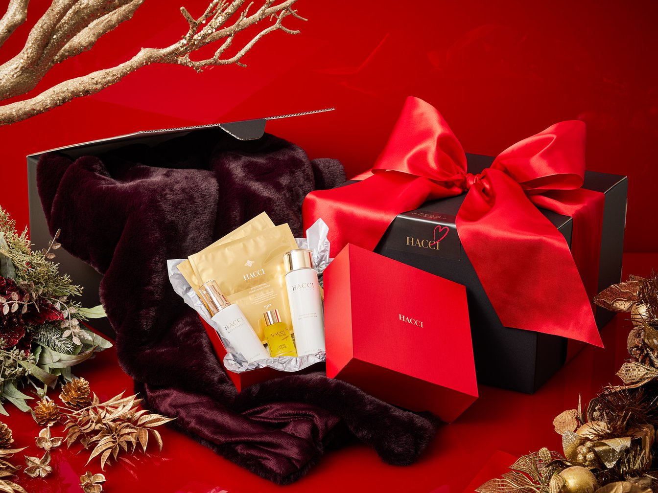 赤いリボンがついたギフトボックスと、ブラウンのブランケットの上に置かれたスキンケアアイテム