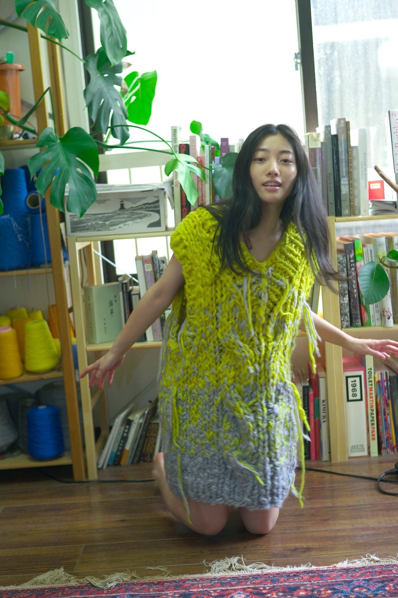 イエローのニットベストを着てアトリエの床に膝立ちする川島鈴遥